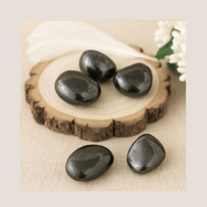 polished Hematite stones on a wooden display block with a white feather and floral accent, showing natural metallic sheen and raw inclusions.