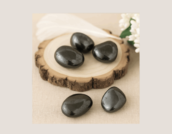 polished Hematite stones on a wooden display block with a white feather and floral accent, showing natural metallic sheen and raw inclusions.
