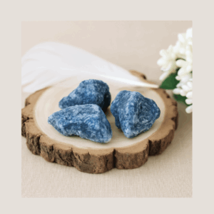 A photograph of three raw Blue Quartz crystals displayed on a wooden block with a white feather and soft flower, showcasing their natural colour, inclusions, and imperfect shapes in the Grounded Goddess crystal image style.