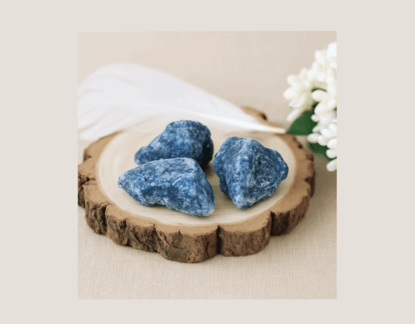 A photograph of three raw Blue Quartz crystals displayed on a wooden block with a white feather and soft flower, showcasing their natural colour, inclusions, and imperfect shapes in the Grounded Goddess crystal image style.